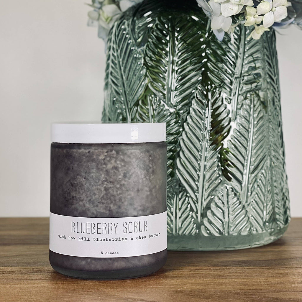Candle labeled 'Blueberry Scrub' in front of a decorative vase with floral arrangement