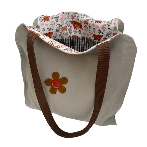 A canvas tote bag with a beige exterior, brown webbed handles, and a floral-bison lining. A brown and orange flower applique is stitched on the front.