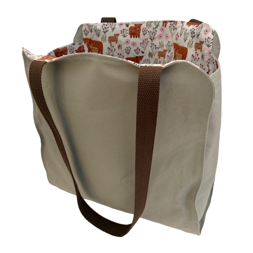 A canvas tote bag with a beige exterior, brown webbed shoulder straps, and a patterned fabric lining featuring brown cows and pink wildflowers on a white background.