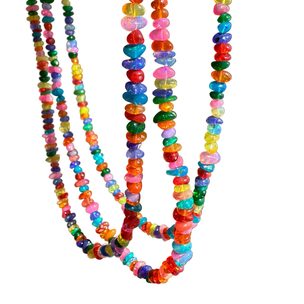 Three strands of colorful beaded necklaces made of translucent, faceted pebble-shaped beads in various bright colors, hanging against a plain beige wall.