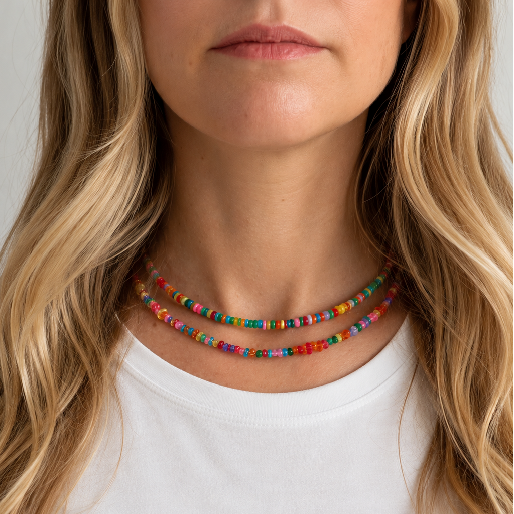 A close-up of a woman wearing a double-strand beaded necklace with colorful, patterned beads. The necklace is shown on her neck above a white t-shirt, with her blonde hair framing the shot against a light gray background.