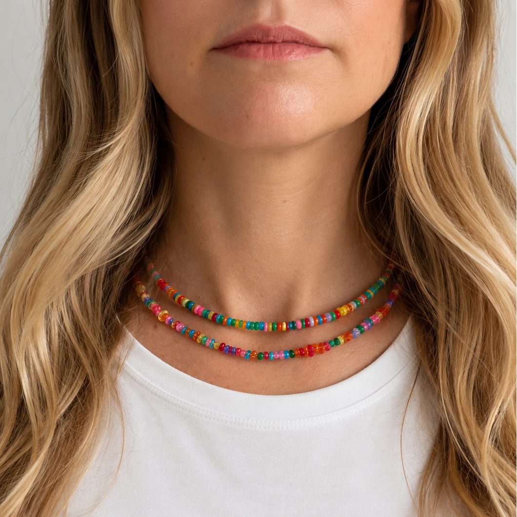 A close-up of a woman wearing a double-strand beaded necklace with colorful, patterned beads. The necklace is shown on her neck above a white t-shirt, with her blonde hair framing the shot against a light gray background.