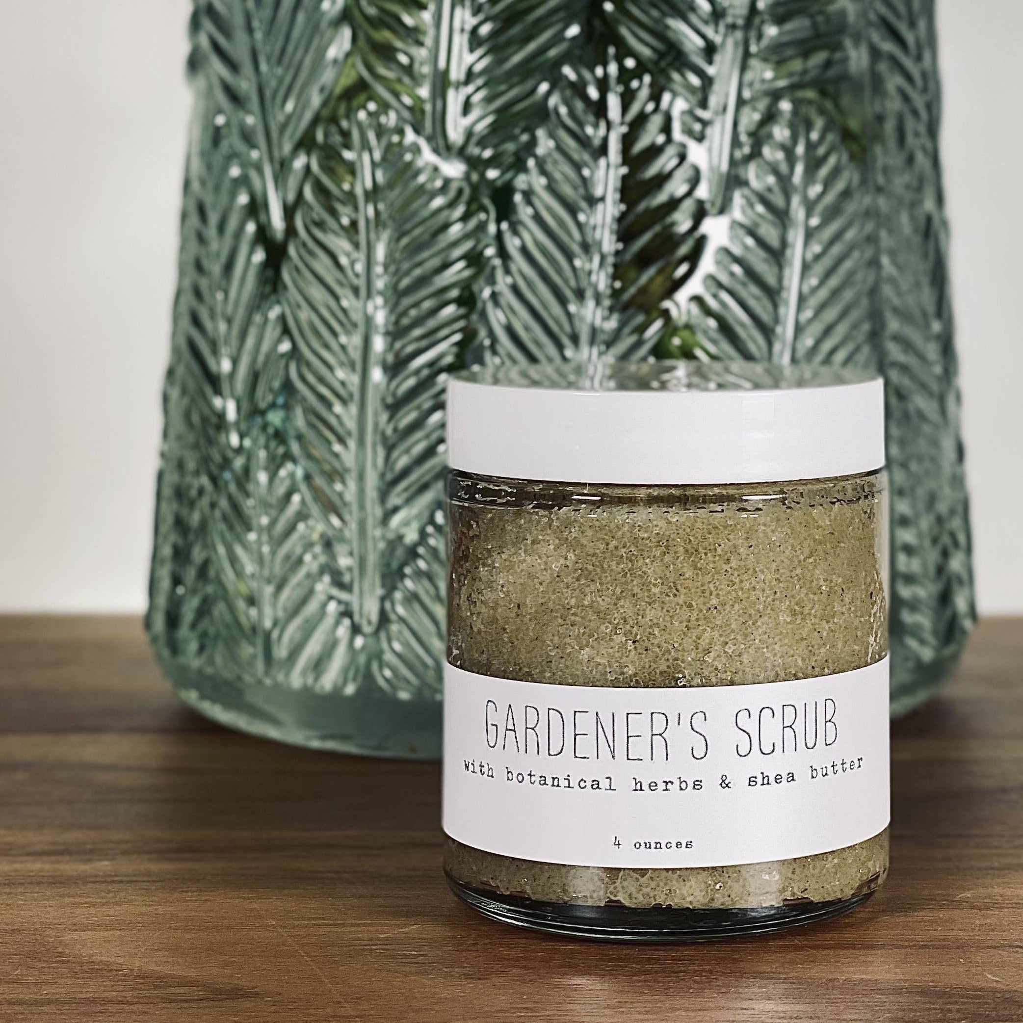 Candle labeled 'Gardener's Scrub' in front of a green vase with flowers