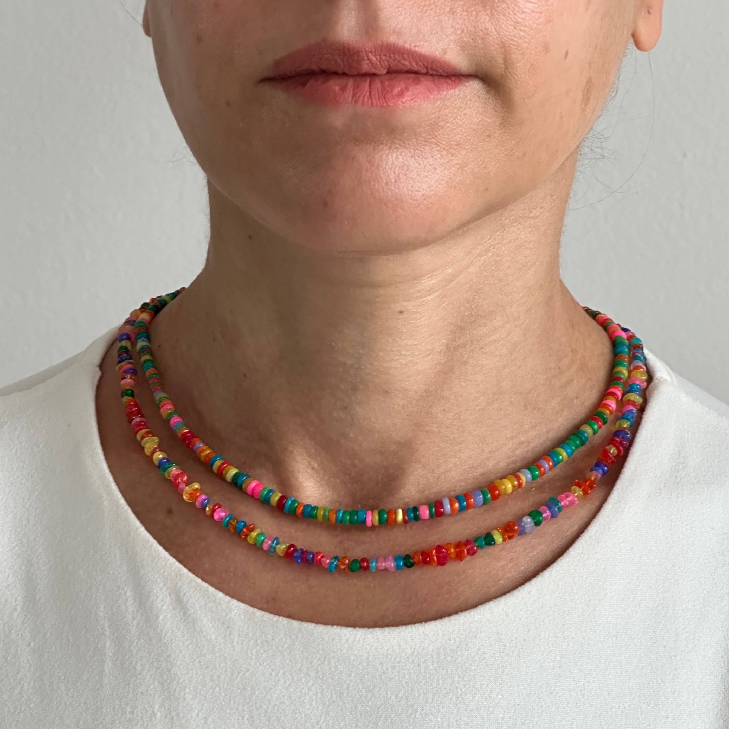 A close-up of a person wearing two layered beaded necklaces with colorful round beads, paired with a white crew-neck top against a plain light gray background.