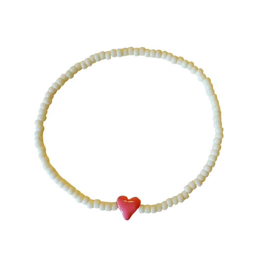 A beaded bracelet made of small off-white round beads with a glossy pink heart-shaped charm at the bottom, shown on a plain white background.