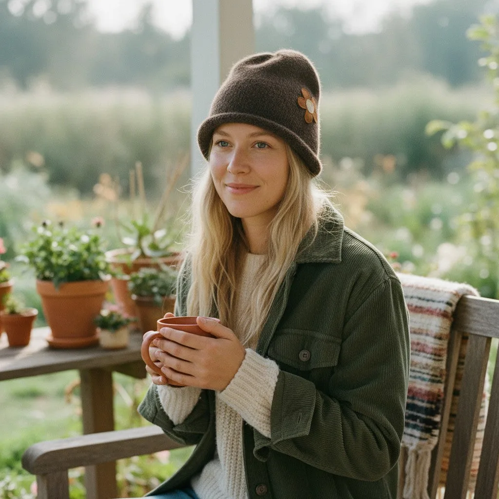 A woman sitting on a wooden porch chair wearing a dark brown knit beanie with a small flower accent, an olive green jacket, and a cream knit sweater, while holding a brown ceramic mug. The background shows a softly blurred garden with potted plants.