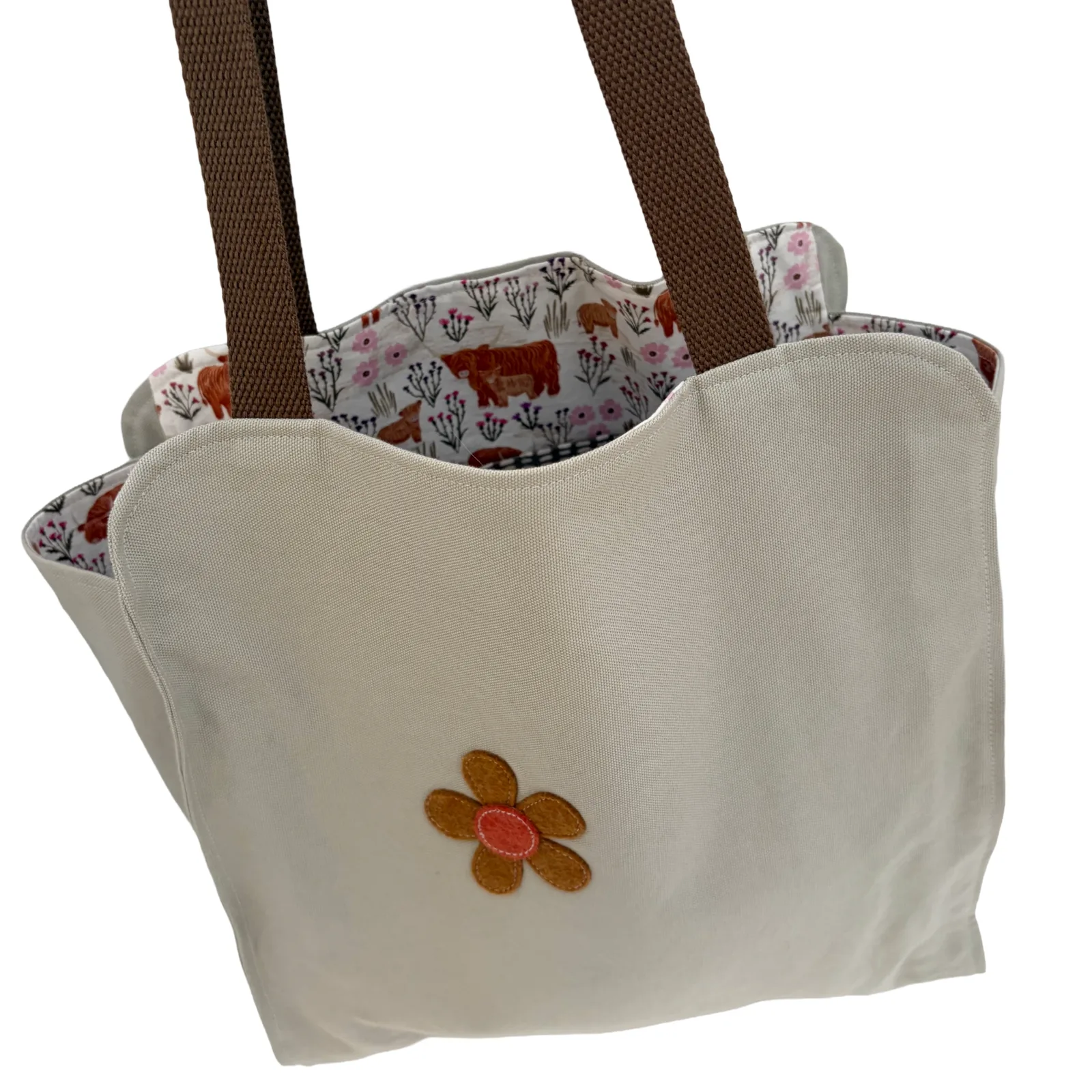 An off-white canvas tote bag with brown shoulder straps, featuring a small embroidered flower on the front and a floral-and-animal print lining the interior.
