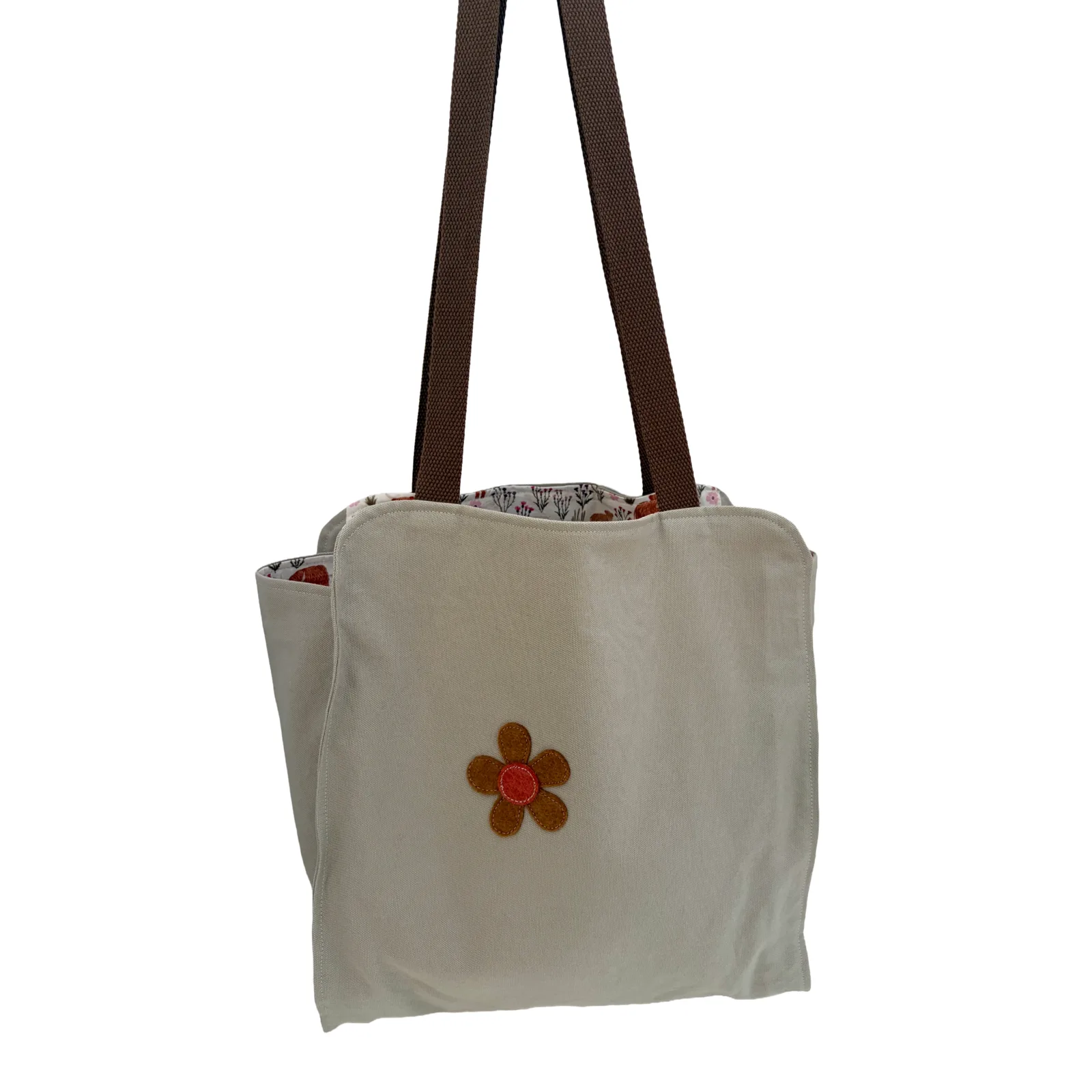 An off-white canvas tote bag with dark brown shoulder straps and a small brown flower applique with a red center on the front, shown against a plain white background.