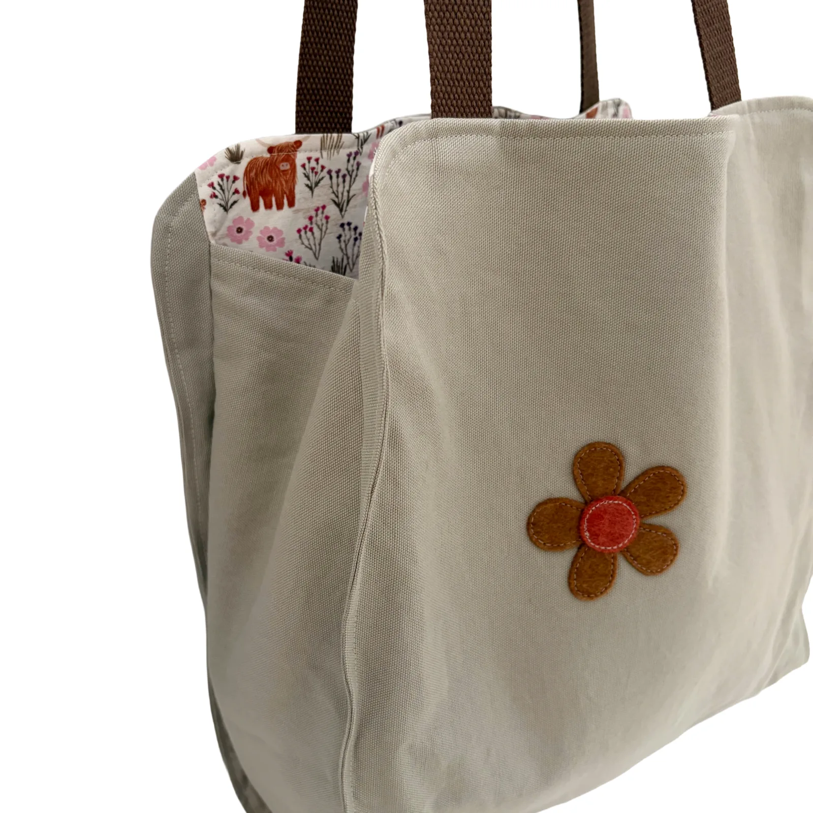 A close-up of a beige canvas tote bag with brown shoulder straps, a floral fabric lining visible in the side pocket, and a brown felt flower applique with a red center on the front.