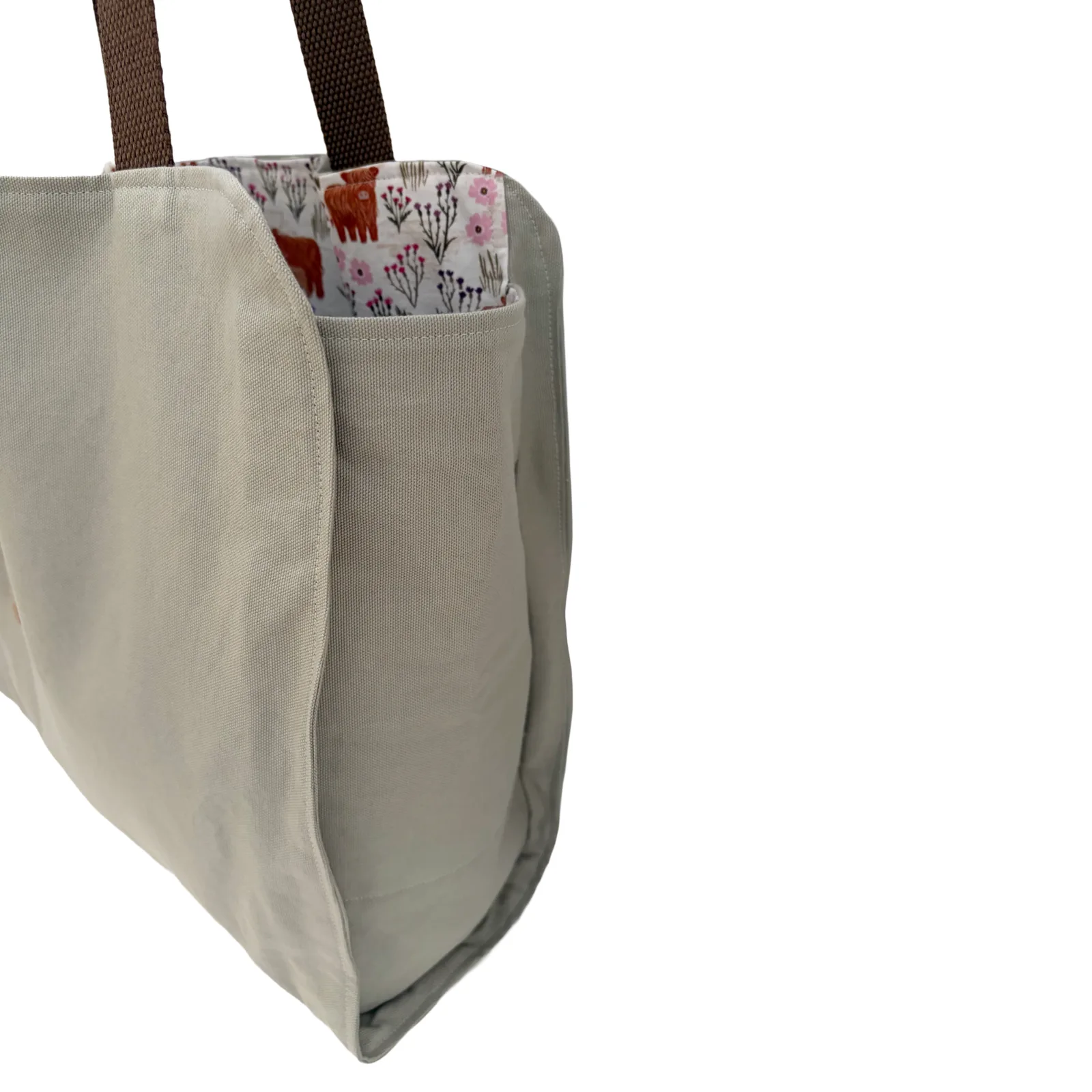 A close-up of a light beige canvas tote bag with dark brown shoulder straps and a floral-patterned fabric lining visible in the side pocket, set against a plain white background.