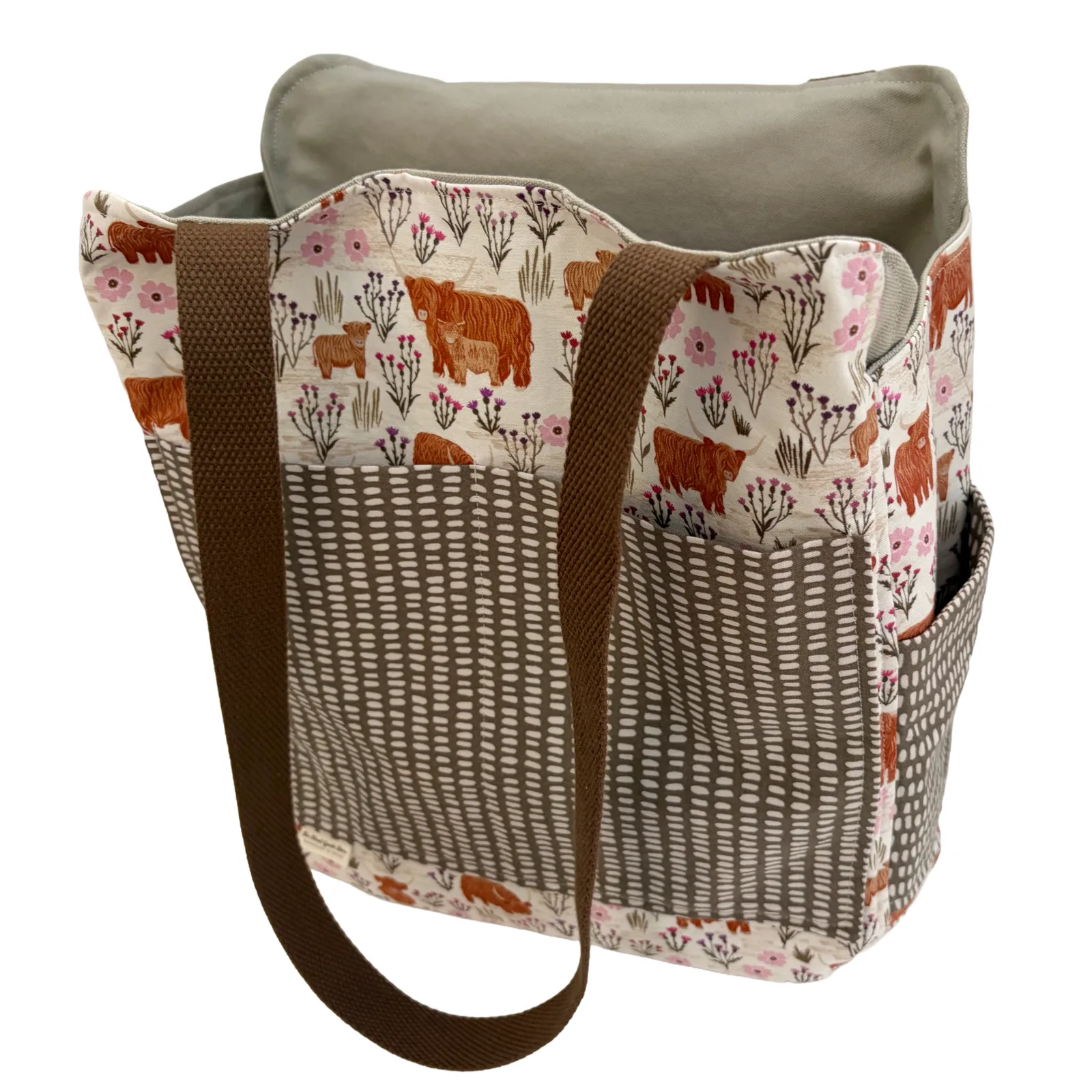 A fabric tote bag with a patchwork design featuring a cream background with brown Highland cows, pink flowers, and a brown woven-texture pattern, with solid brown handles and a matching brown interior.