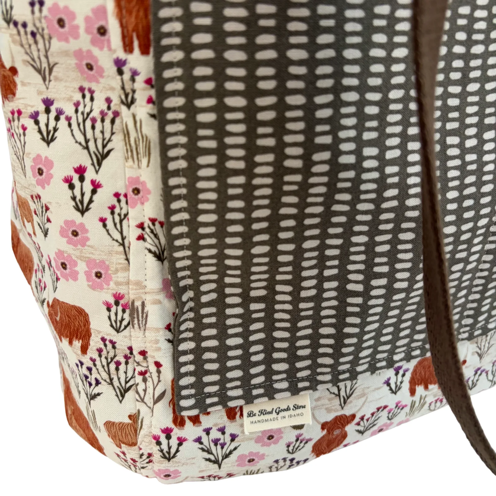 Close-up of a fabric tote bag corner showing a cream print with pink wildflowers, brown deer, and a dark gray patterned panel with a brown strap.