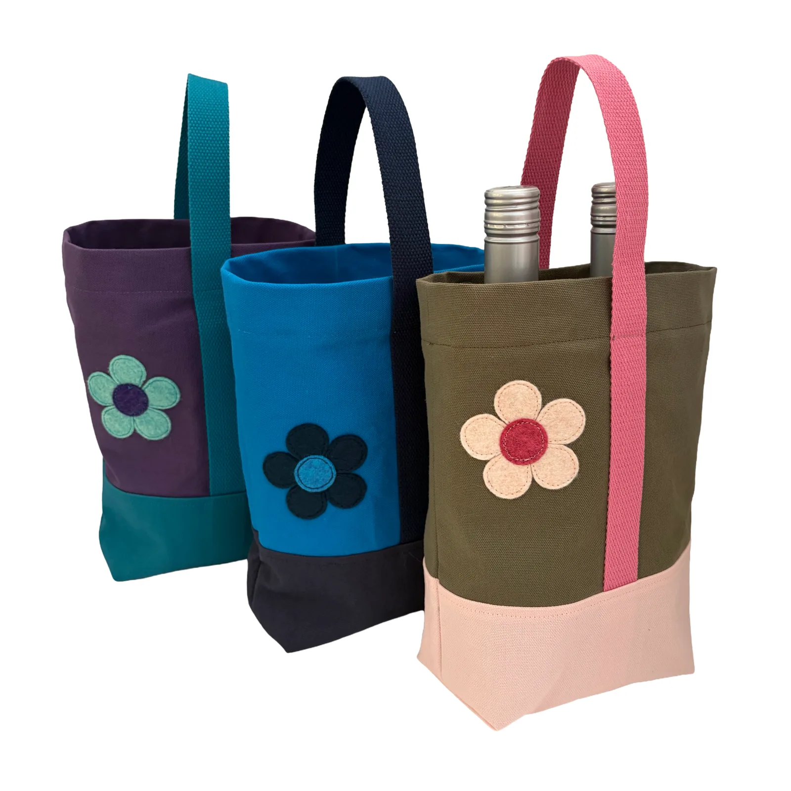 Three fabric wine bottle bags with floral appliques, shown on a white background. The bags are olive green with a pink handle and base, blue with a black handle and base, and purple with a teal handle and base.