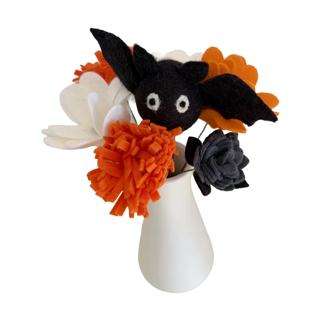 A Halloween-themed felt flower bouquet in a small white vase, featuring a black bat with white eyes, orange pom-poms, a white flower, and a dark gray felt rose.