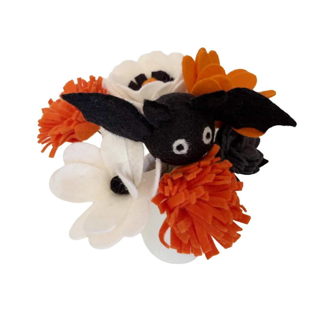 A Halloween-themed felt hair clip featuring a black felt bat with white eyes, surrounded by orange and white felt flowers and pom-poms, isolated on a white background.