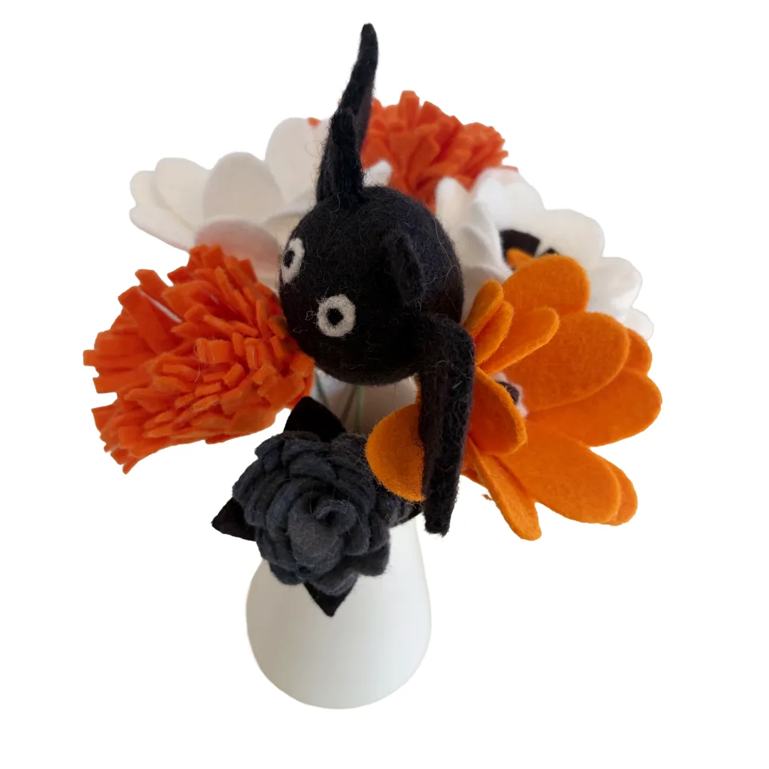A small black felt bat decoration with white eyes, nestled in a floral arrangement of orange, white, and dark gray felt flowers on a white vase against a white background.
