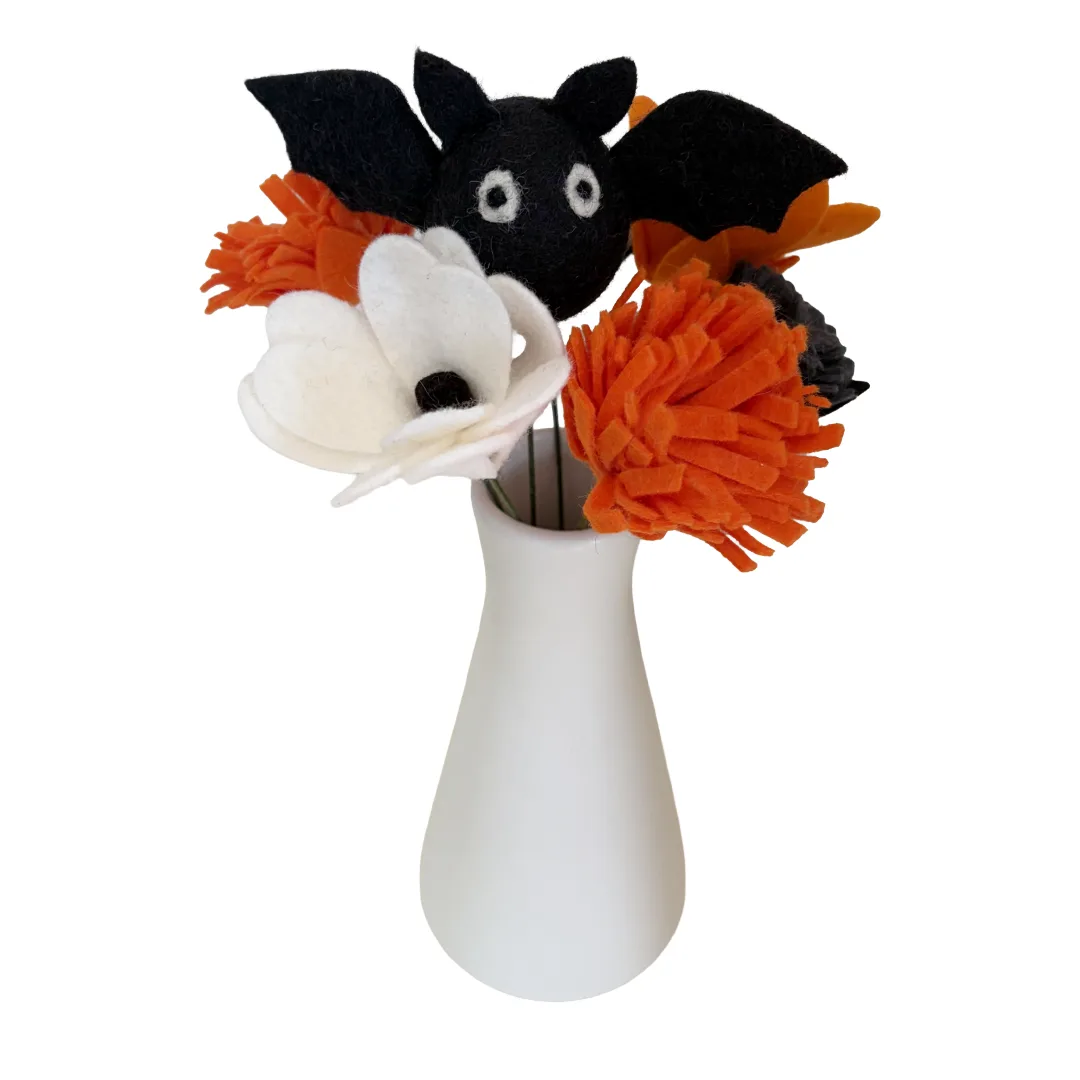 A Halloween-themed felt flower arrangement in a small white vase, featuring a black bat, orange pom-poms, and a white flower with a black center.
