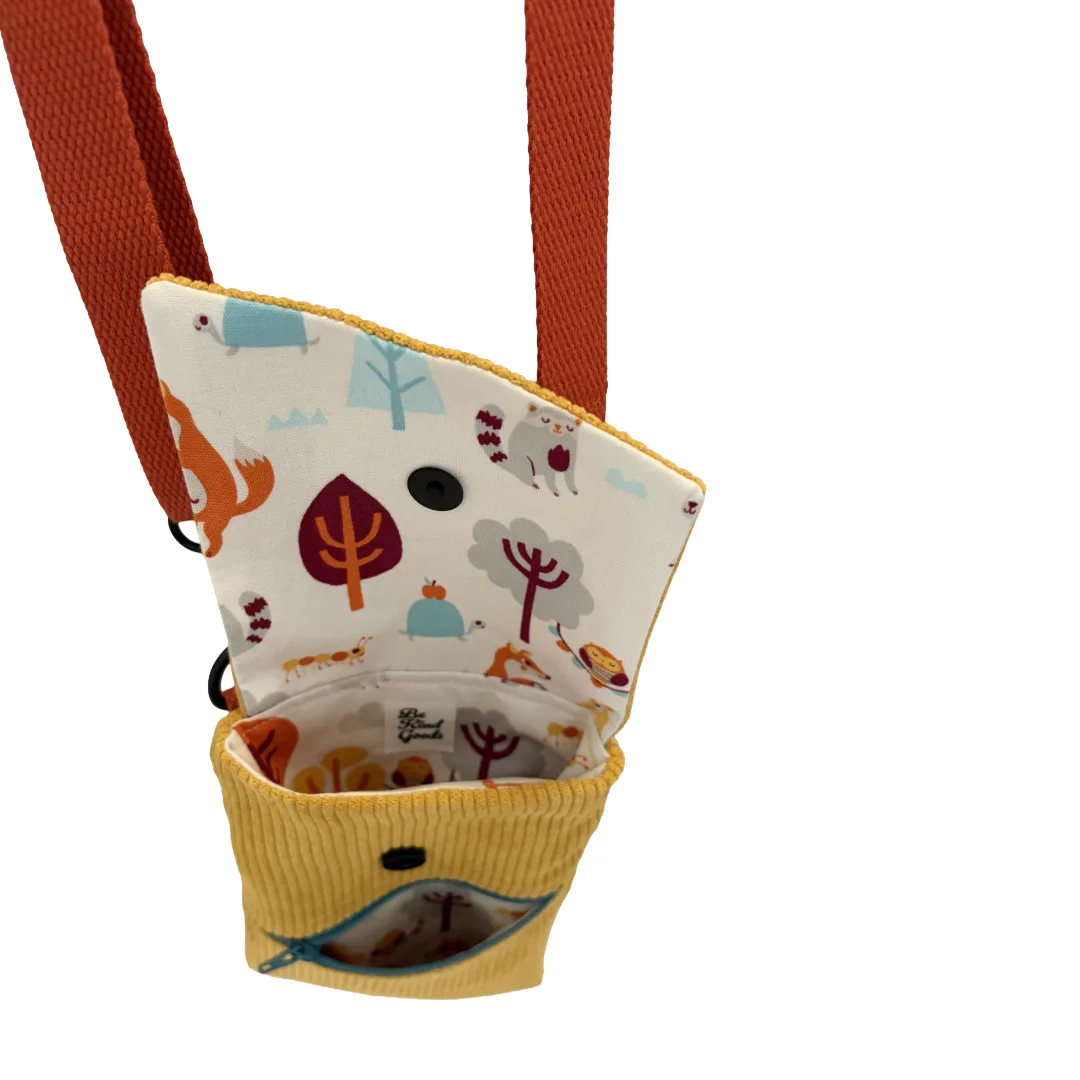 A small fabric pouch with a wide orange-brown shoulder strap, shown open against a plain white background. The pouch has a yellow ribbed fabric front pocket and a white printed interior and flap featuring woodland illustrations such as trees, animals, and small objects.