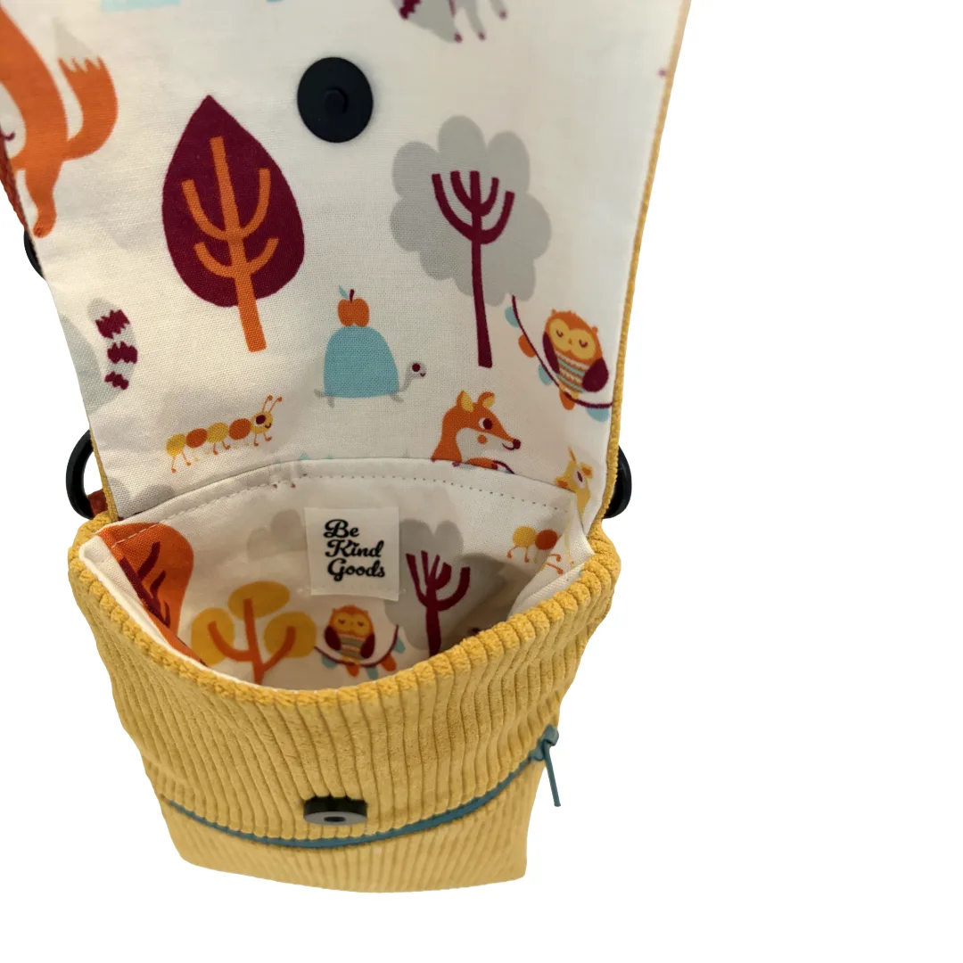 A close-up of a fabric pouch or small bag with a yellow corduroy lower section and a white upper flap printed with colorful woodland animals and trees. The interior label reads 'Be Good Goods'.