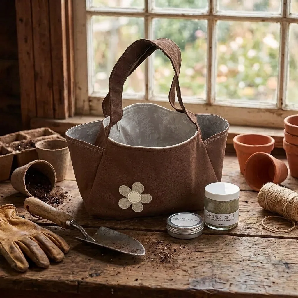A brown canvas gardening tote bag with a silver metal liner and a white flower applique, displayed on a rustic wooden table with gardening tools, pots, and a window in the background.