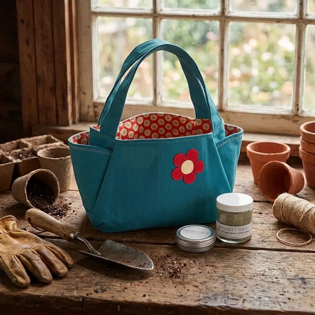 A teal fabric gardening tote bag with a red flower applique sits on a rustic wooden table surrounded by gardening tools, including gloves, a trowel, terracotta pots, twine, and small jars, with a window in the background.