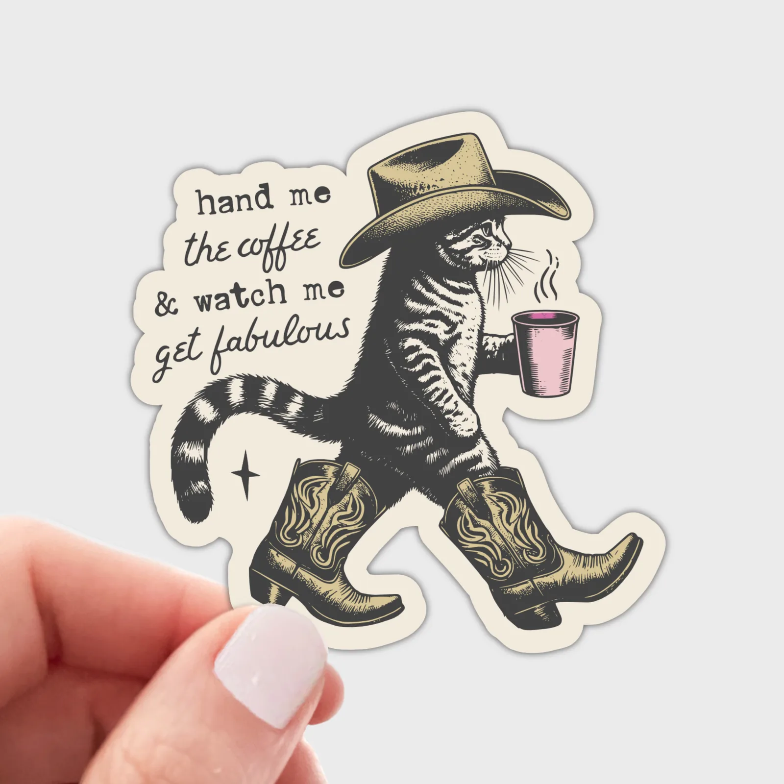 A die-cut sticker featuring a walking tabby cat in a cowboy hat and boots, holding a pink coffee cup, with the text 'hand me the coffee & watch me get fabulous.' A human hand with pale pink nail polish is holding the sticker against a light gray background.