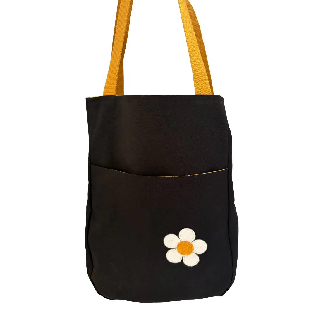 A black canvas tote bag with mustard yellow shoulder straps and a white daisy patch on the front pocket, shown against a plain white background.