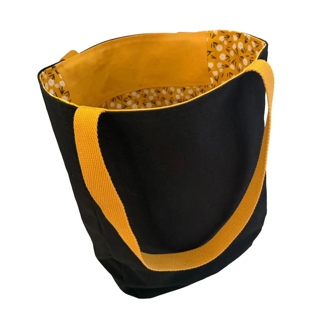 A black canvas tote bag with a mustard yellow handle and a matching mustard yellow interior lining featuring a small white floral pattern.