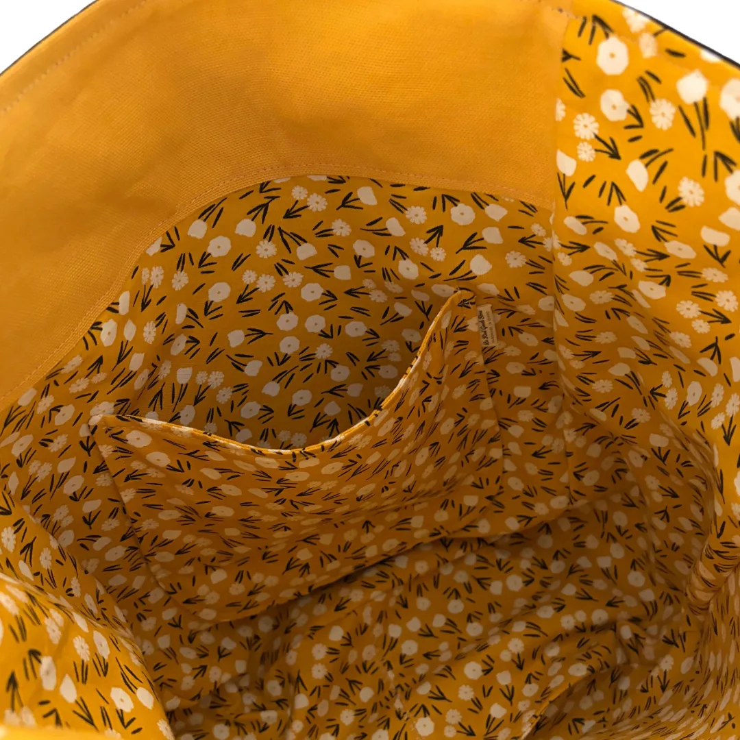 Close-up of the interior lining of a yellow tote bag, featuring a floral and leaf print pattern in white and black, with a solid yellow fabric rim.