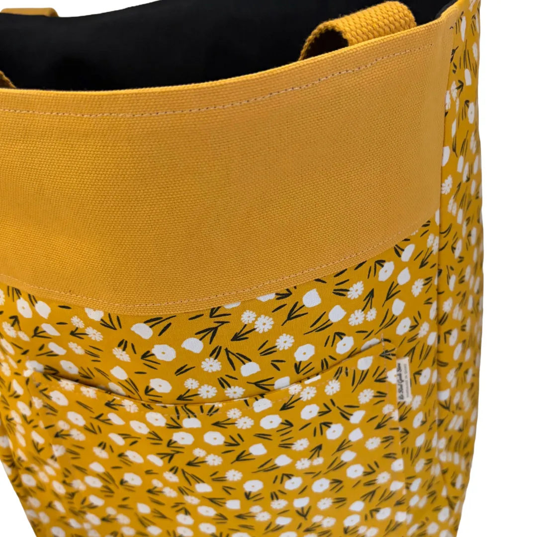 Close-up of a tote bag with a wide mustard yellow top band and a yellow body covered in a white and black floral pattern. The black fabric lining and woven handles are visible at the top.