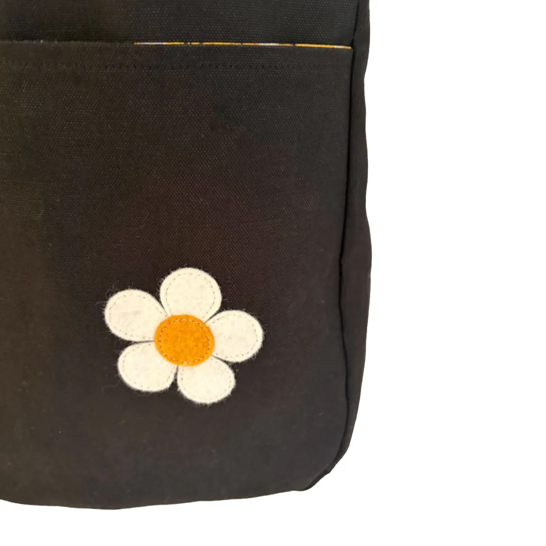 Close-up of a dark brown canvas bag featuring a white and orange embroidered daisy patch on the front pocket.