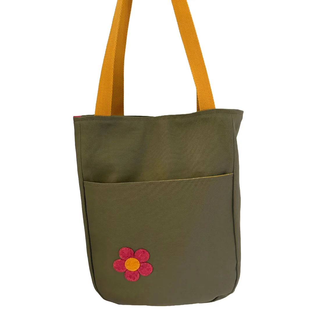 An olive green tote bag with mustard yellow shoulder straps and a small embroidered pink and yellow flower on the front pocket, shown against a plain white background.