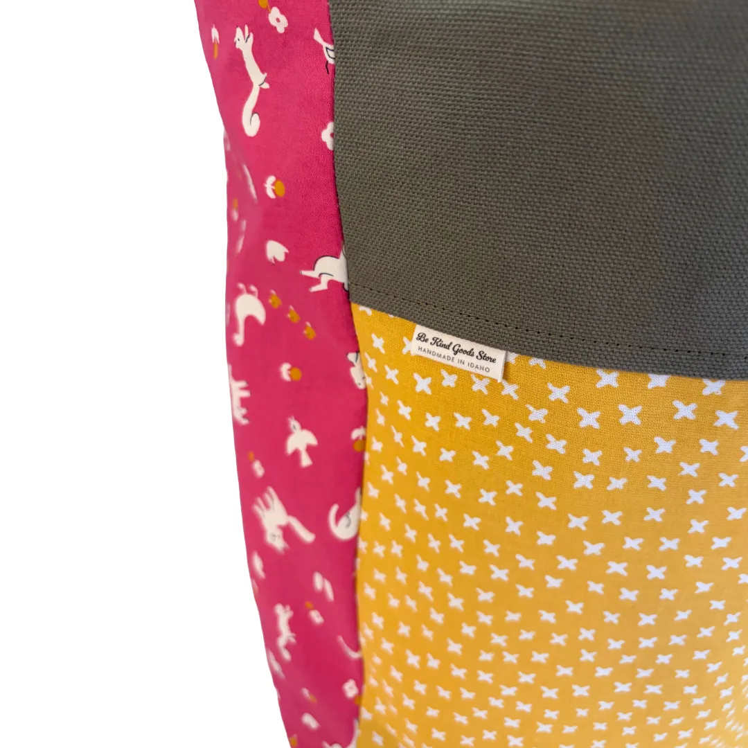 Close-up of a patchwork fabric item, likely a pouch or bag, showing three color-blocked panels: a dark olive canvas section, a mustard yellow panel with white star patterns, and a hot pink panel with white animal silhouettes. A small sewn-on label reads 'Be Kind Goods Store'.