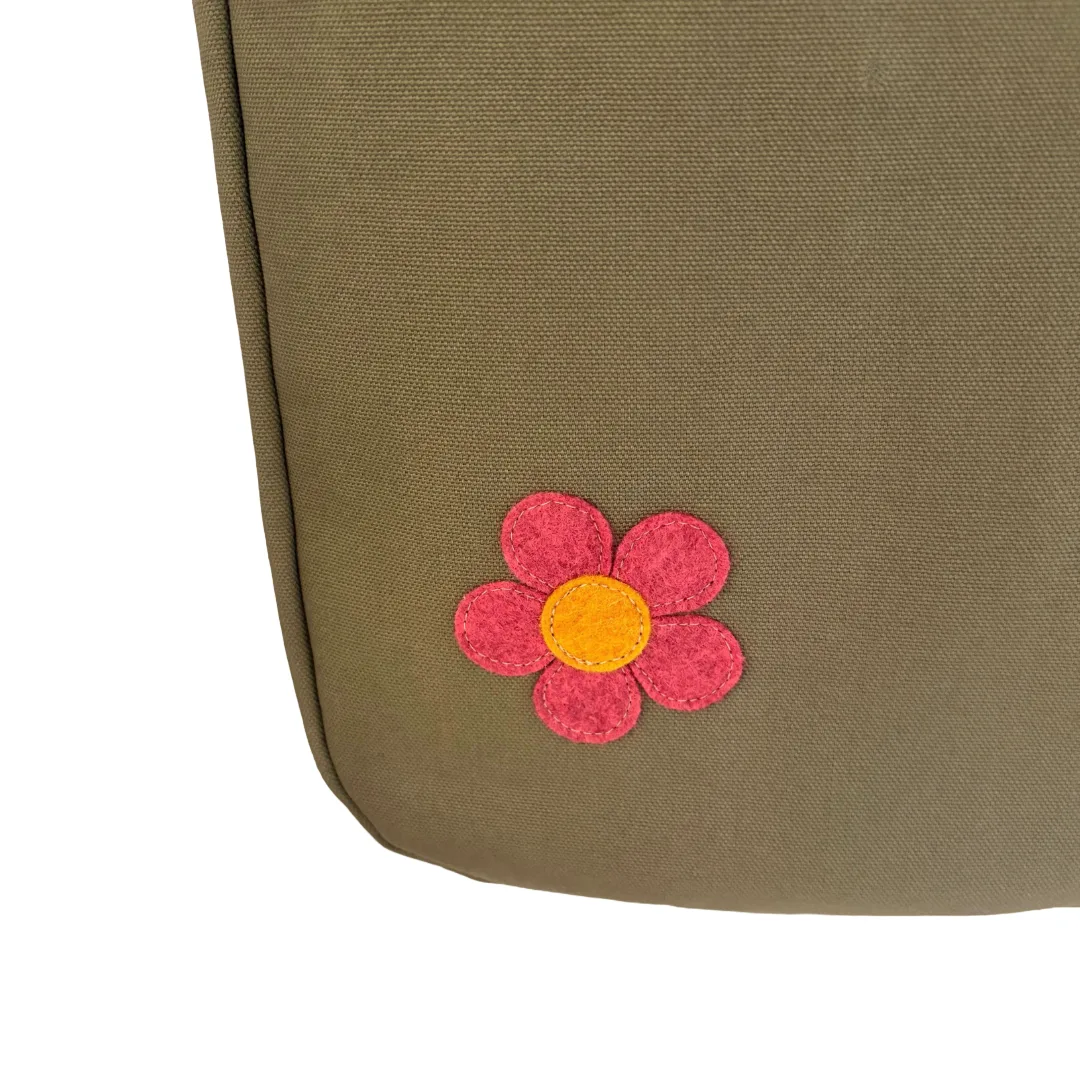 Close-up of an olive green canvas bag featuring a small pink felt flower applique with a yellow center on the lower right corner, set against a plain white background.