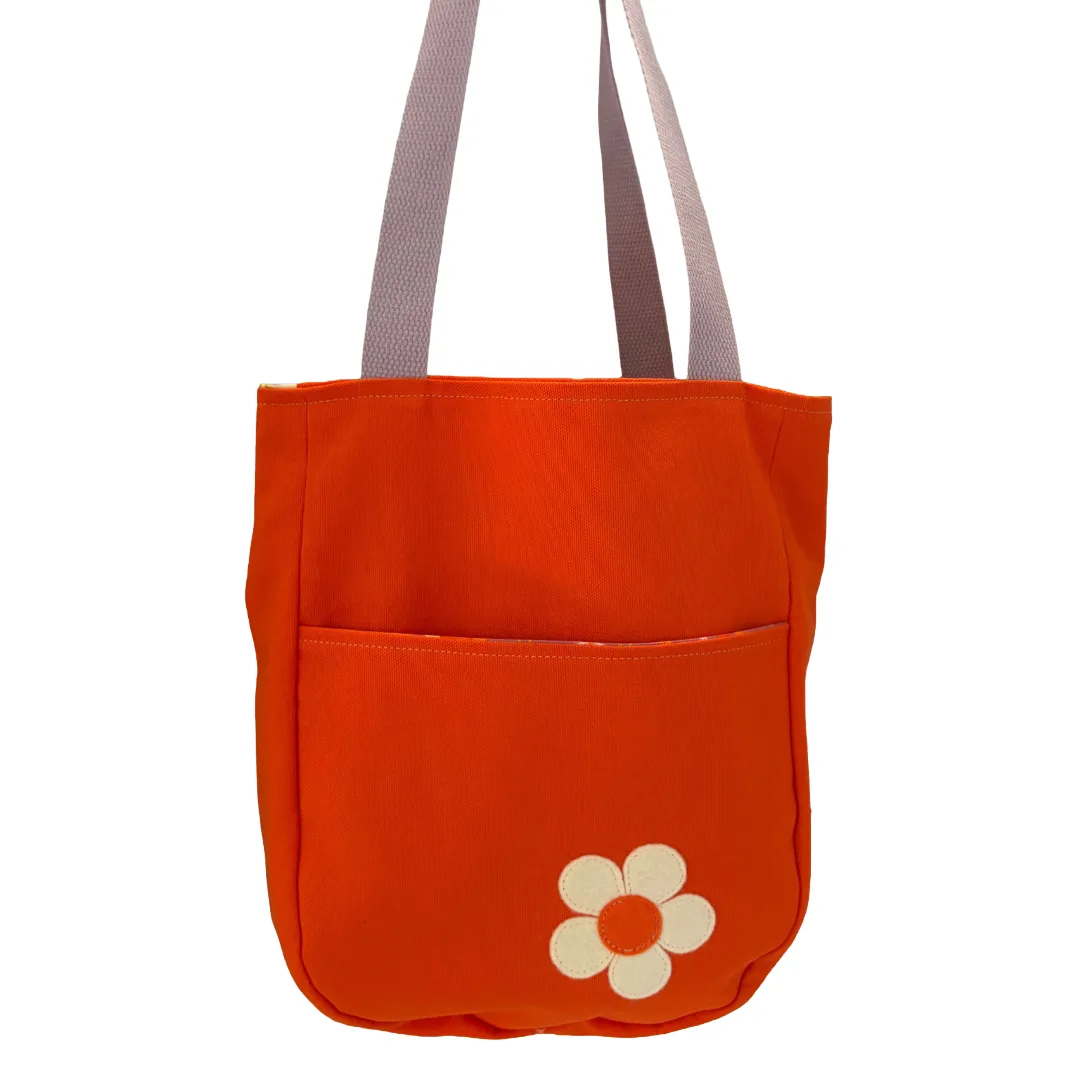 An orange canvas tote bag with a front pocket featuring a cream-colored flower applique, and light gray shoulder straps, shown against a plain white background.