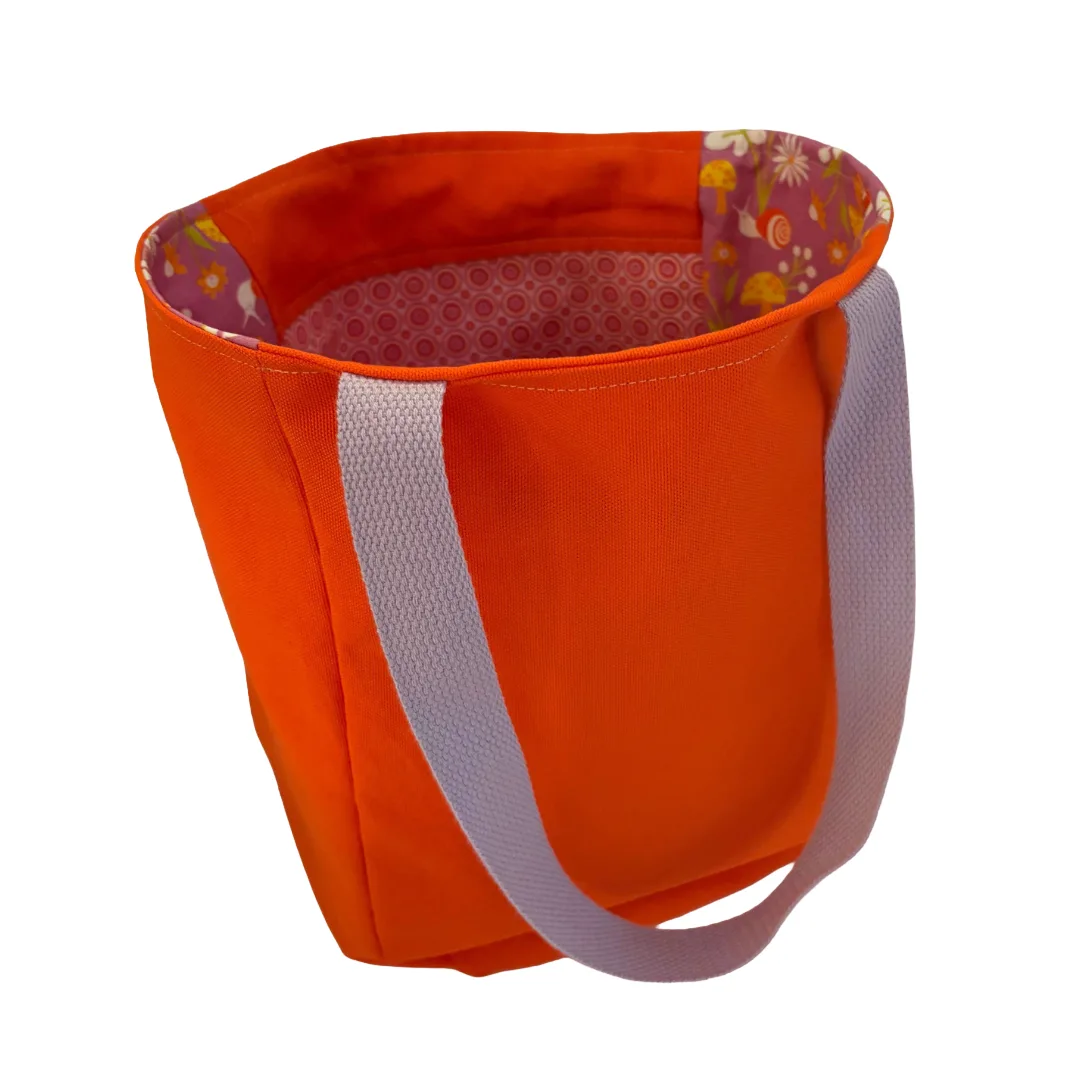 An orange canvas tote bag with light gray fabric handles and a colorful floral pattern lining the interior, shown on a plain white background.
