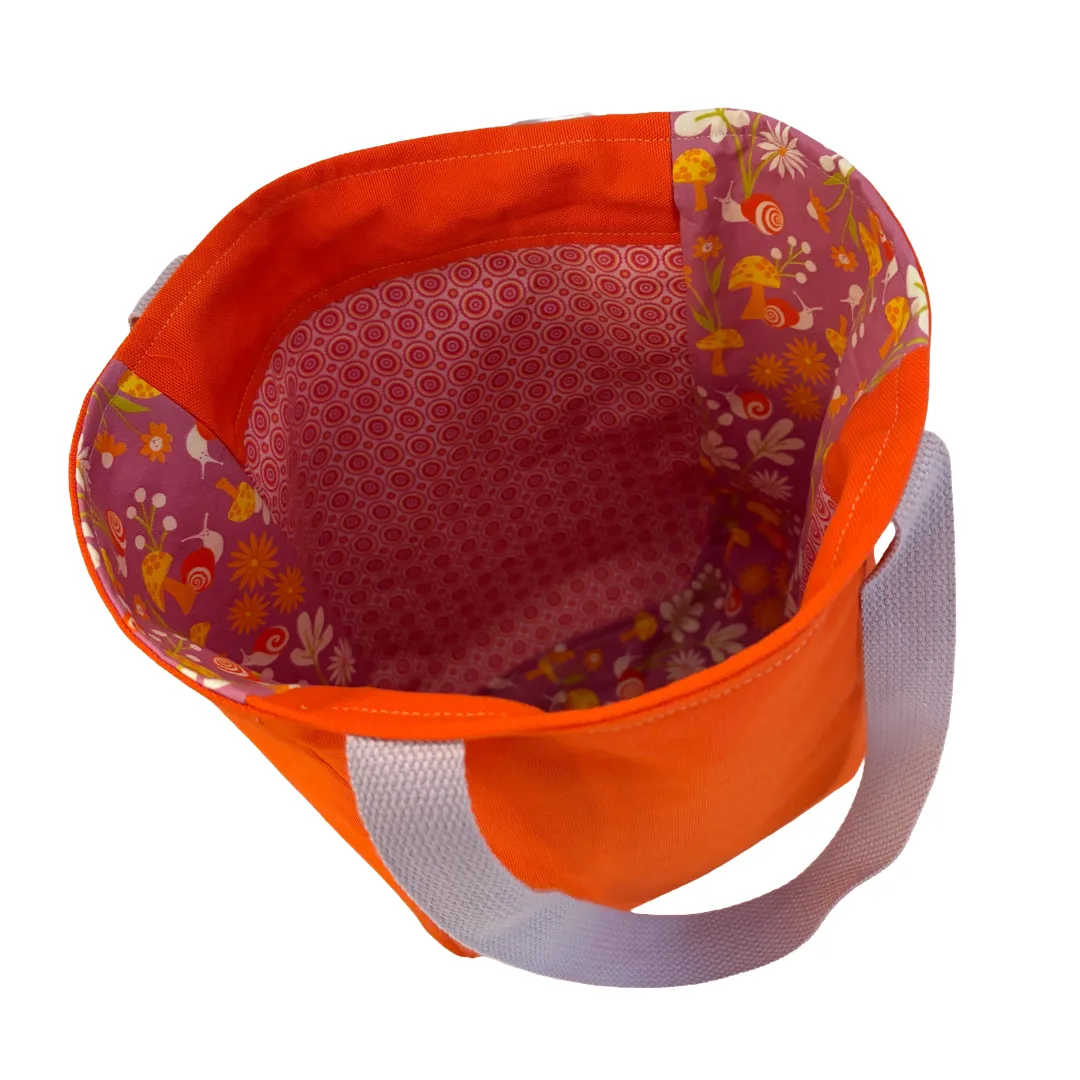 An orange fabric tote bag shown from an overhead angle, featuring a floral patterned interior lining and a light gray webbed handle on a white background.