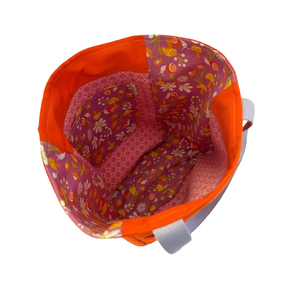 An open fabric bag viewed from above, showing a bright orange exterior and a floral patterned interior in shades of pink, red, yellow, and white. A light gray strap is visible on the right side against a plain white background.