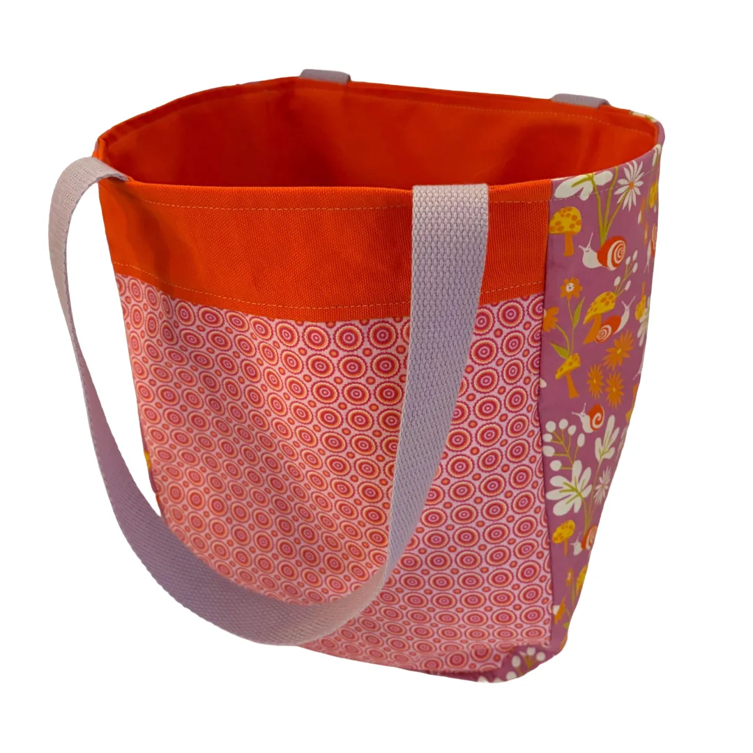A fabric tote bag with a bright orange interior and top trim, a pink and white circular patterned front panel, a purple side panel with yellow floral and butterfly motifs, and light gray fabric shoulder straps.