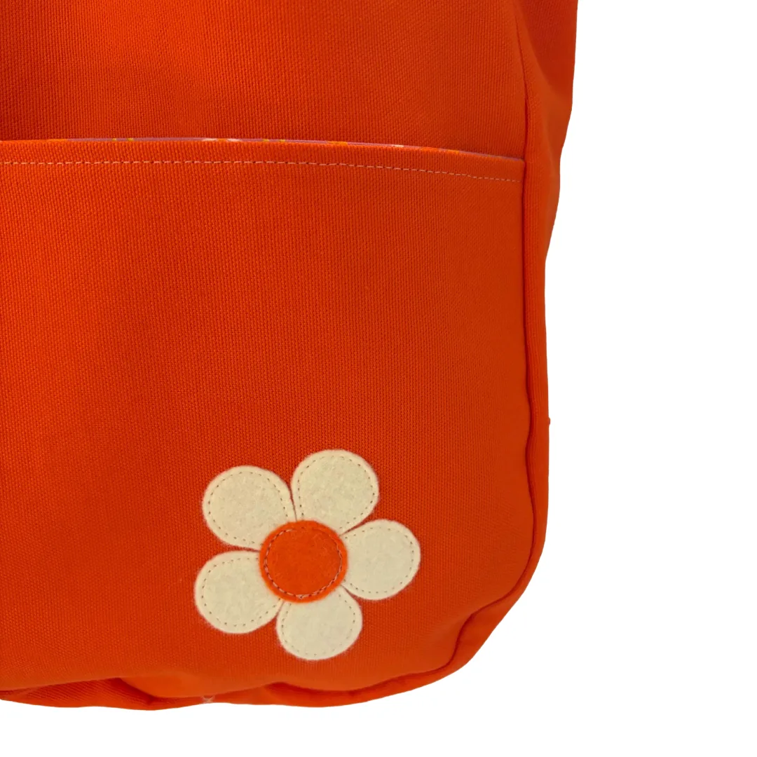 Close-up of a bright orange fabric item, likely a backpack or bag, featuring a cream-colored felt flower applique with an orange center on the lower front pocket.