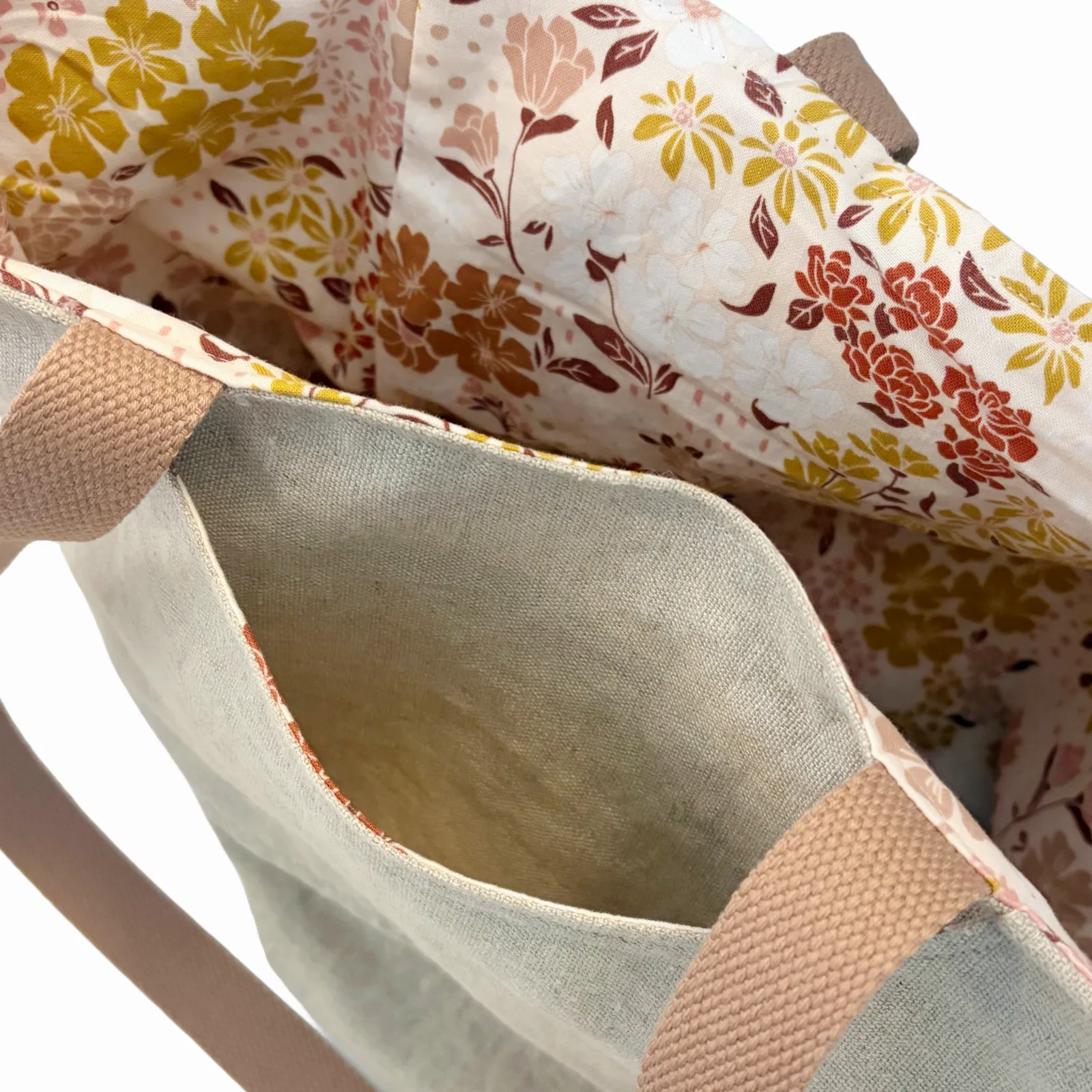 Close-up of a tote bag's interior, showing a light beige canvas exterior, a floral patterned lining in yellow, orange, and brown, and tan webbing handles.