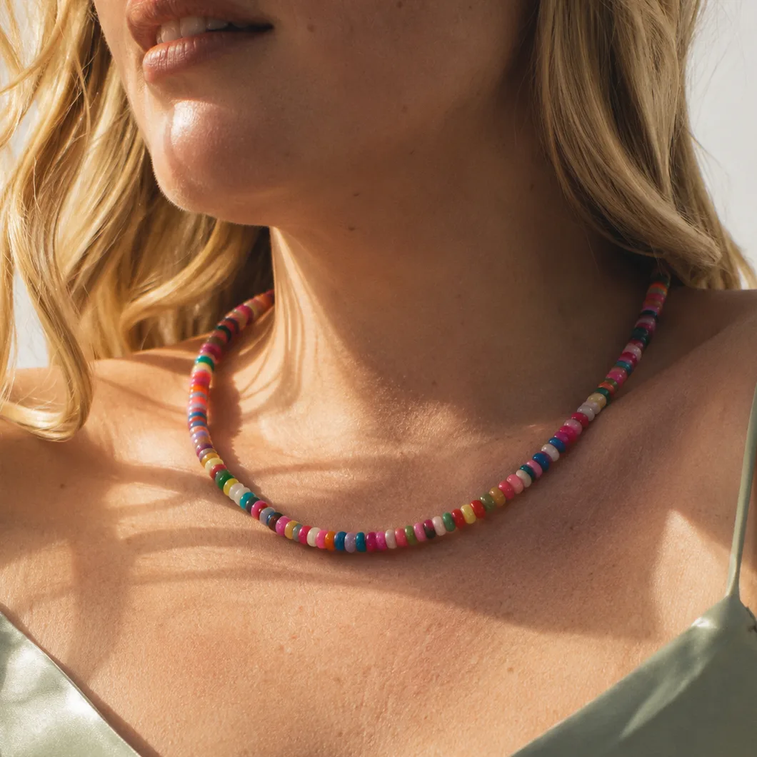 A close-up lifestyle shot of a woman wearing a colorful beaded necklace. The necklace features small, round beads in multiple vibrant colors including pink, blue, yellow, green, and orange.