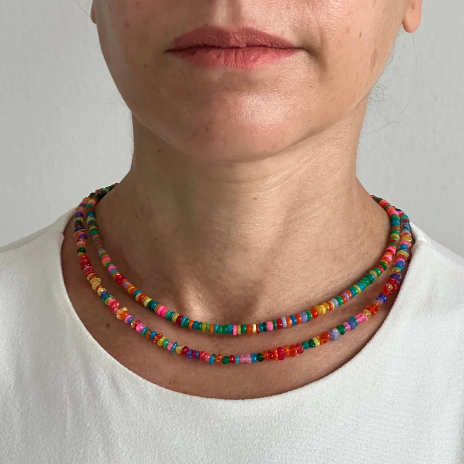 A close-up of a person wearing two layered beaded necklaces with colorful round beads, paired with a white crew-neck top against a plain light gray background.