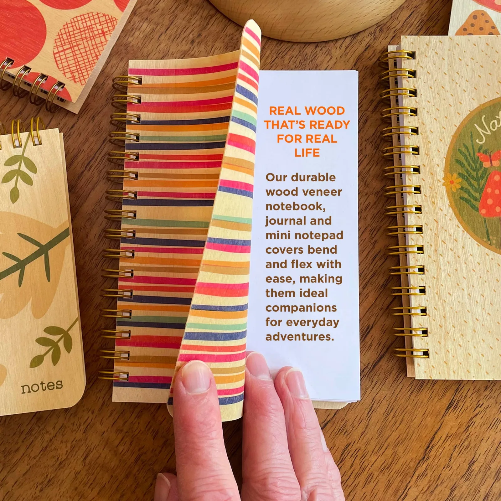 A hand holds a colorful striped spiral notebook in the center of a wooden table, surrounded by other wood-veneer notebooks. The center notebook has a white cover with orange text reading 'REAL WOOD THAT'S READY FOR REAL LIFE'.