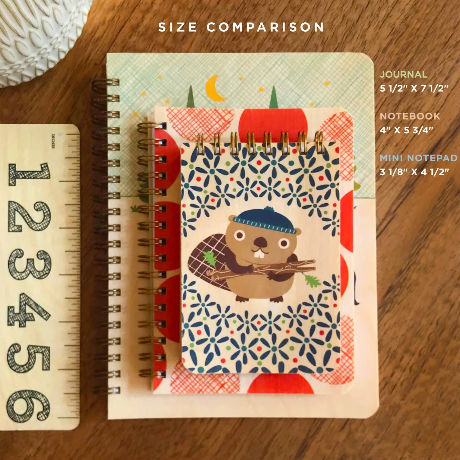 Size comparison of a journal, notebook, and mini notepad with a ruler on a wooden surface.