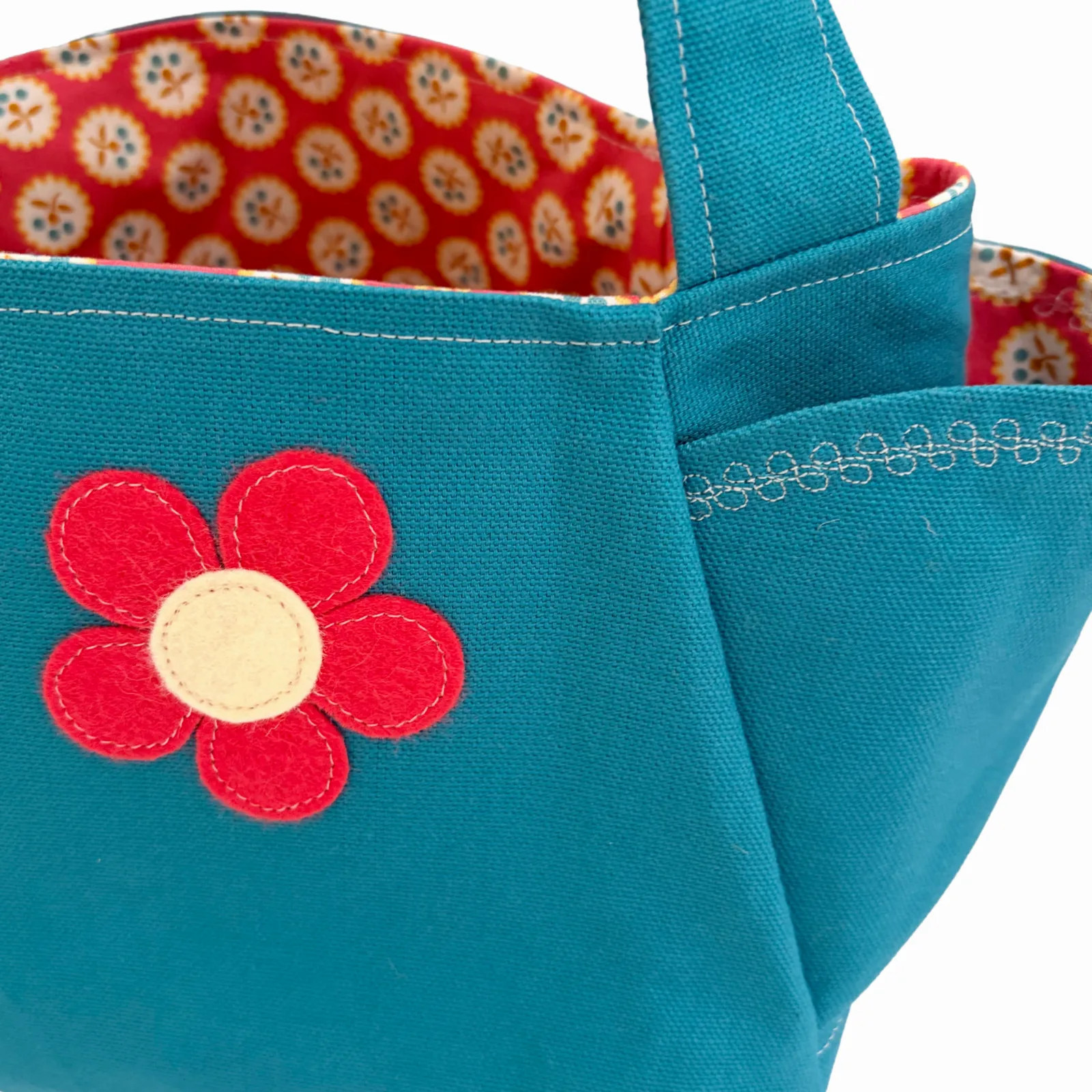 Close-up of a teal canvas tote bag with a red felt flower applique on the front, white contrast stitching, and a red patterned fabric lining visible inside.