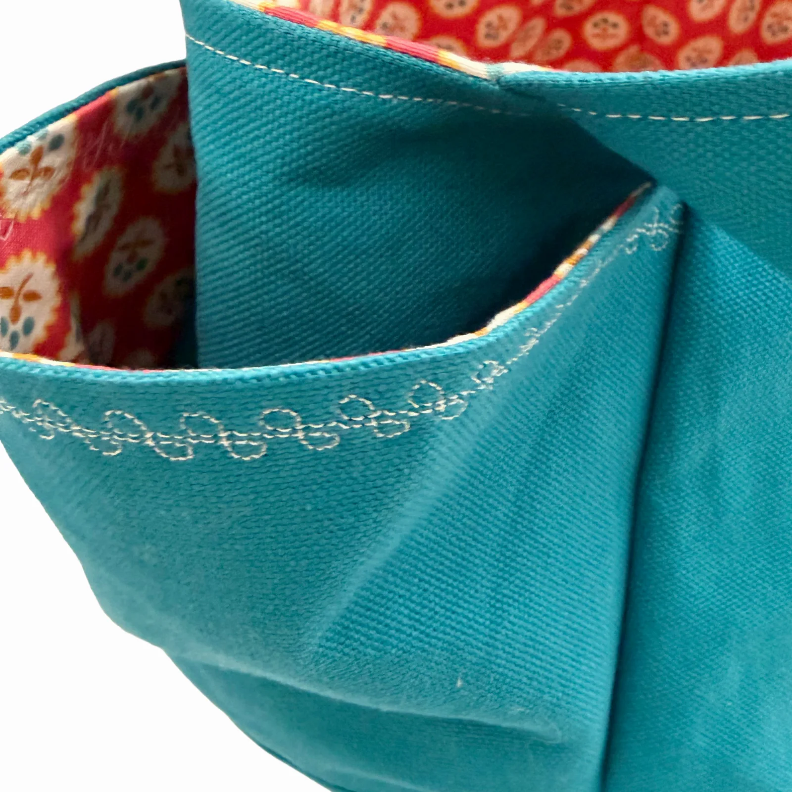 Close-up of a teal canvas tote bag with white decorative stitching and a red patterned fabric lining visible inside the pocket.