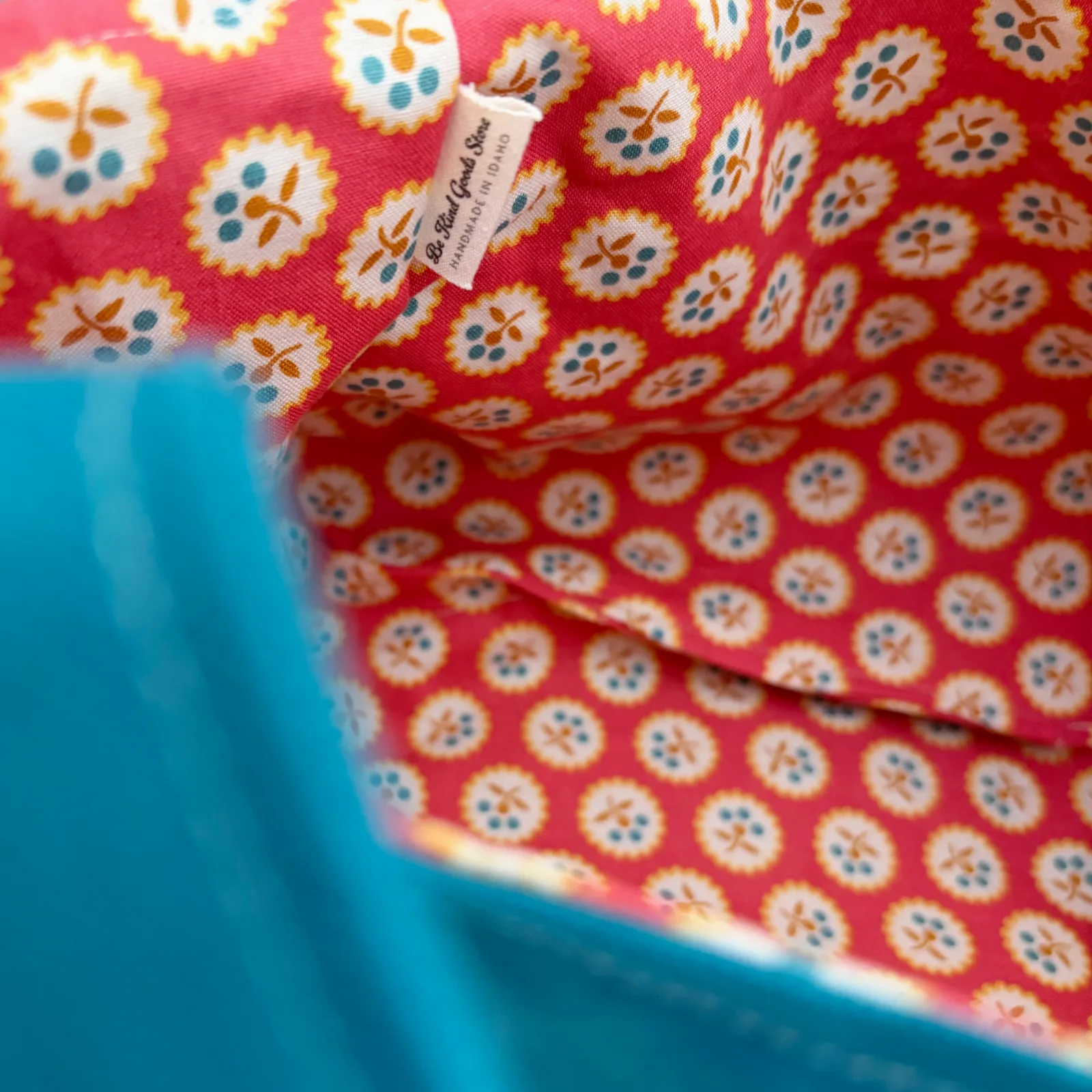 Close-up of a fabric product with a red background and a repeating white, yellow, and blue floral pattern, showing a small sewn label that reads 'The Real Cat Bag'.
