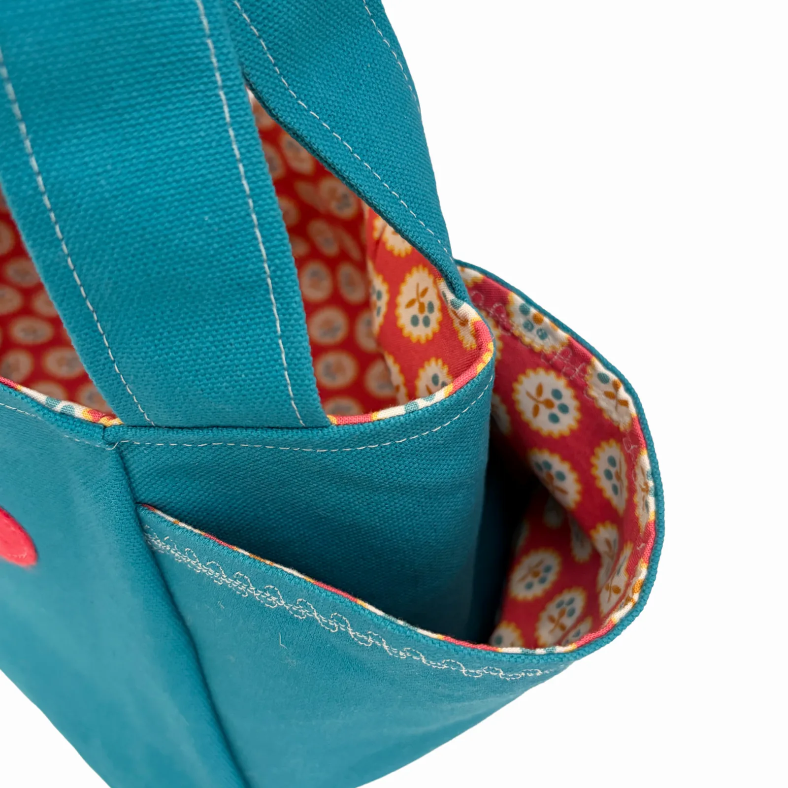 Close-up of a teal canvas tote bag showing the interior lining with a red, orange, and cream floral pattern, white stitching, and a side pocket.