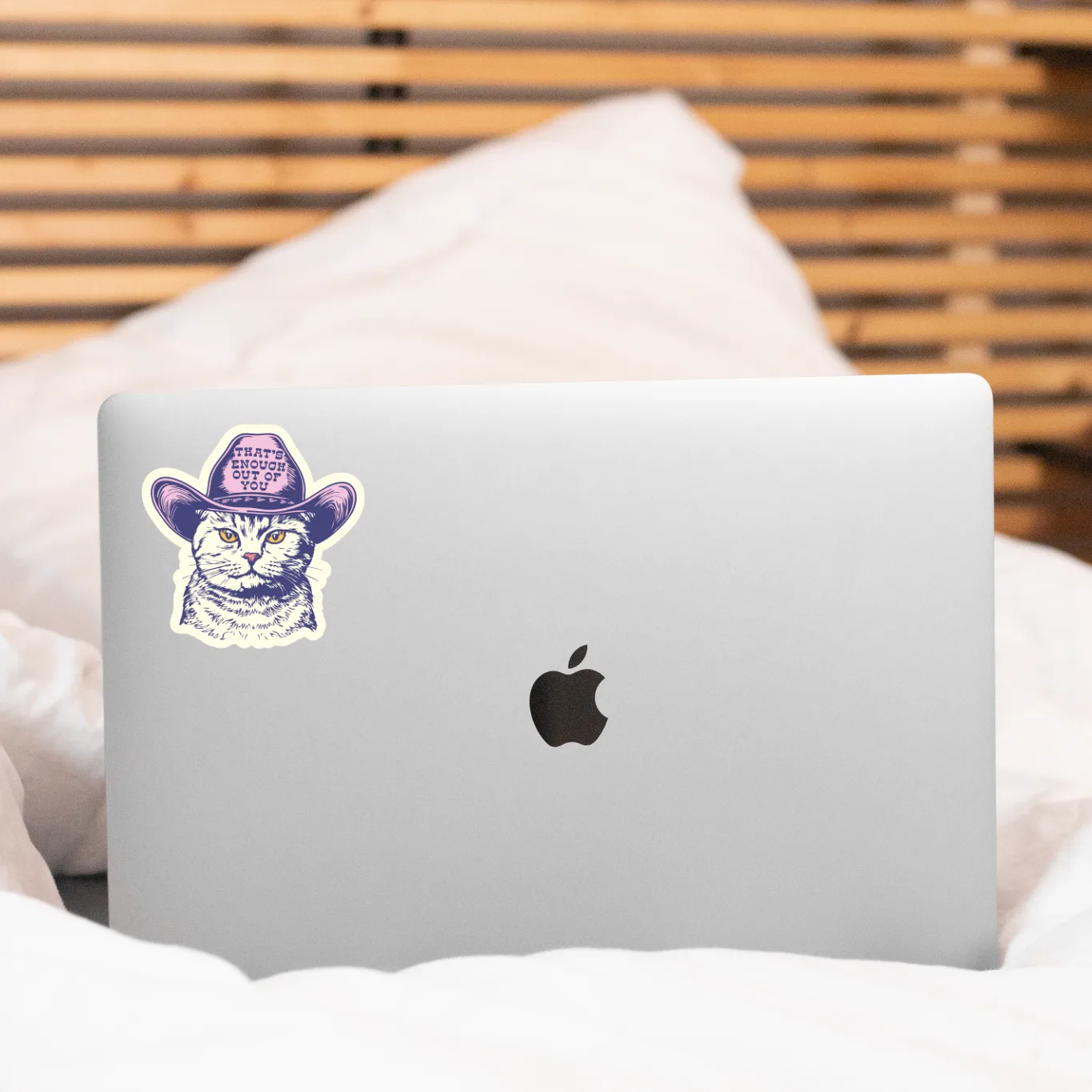 A silver laptop with a purple and white cat sticker wearing a cowboy hat, resting on white bedding in front of a wooden slatted headboard.